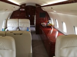 Luxurious inside of a private jet. White leather chairs and mahogany dresser with fruit on top.