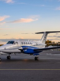 Small, light grey jet with blue engines parked at Oslo Airport Gardermoen. Light blue evening sky with peach clouds in the background
