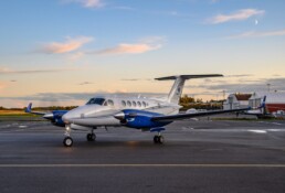 Small, light grey jet with blue engines parked at Oslo Airport Gardermoen. Light blue evening sky with peach clouds in the background