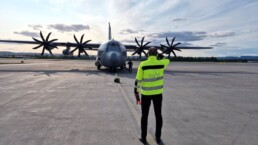 Man in yellow jacket is marshalling a four propelled gray military plane at Gardermoen Airport.